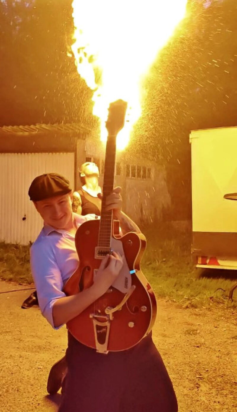 Simon die een gitaar omhoog houd met in de achtergrond een vuurspuwer die vuur uit zijn mond spuugt